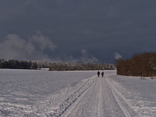 istock Beautiful winter landscape with two elderly people walking on snow-covered path on sunny day near Burladingen, Swabian Alb, Germany with forest on the horizon. 1295821996