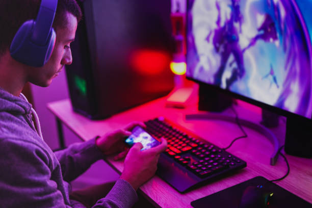 Young boy wearing headset and play video games with smartphone online in front of computer - Home isolated for coronavirus outbreak Young boy wearing headset and play video games with smartphone online in front of computer - Home isolated for coronavirus outbreak playing alone stock pictures, royalty-free photos & images