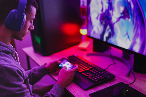 молодой мальчик носить гарнитуру и играть в видеоигры со смартфоном онлайн перед компьютером - главная изолированы от вспышки коронавирус� - gamer стоковые фото и изображения