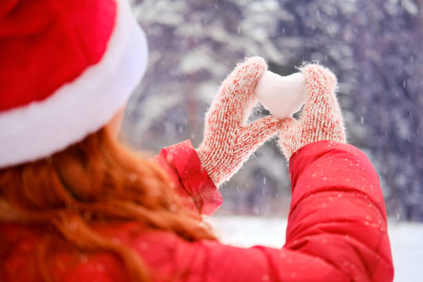 una donna con un cappello di babbo natale tiene un cuore fatto di neve su uno sfondo di alberi della foresta, da vicino. donna in possesso di una palla di neve a forma di cuore per san valentino - glove winter wool touching foto e immagini stock