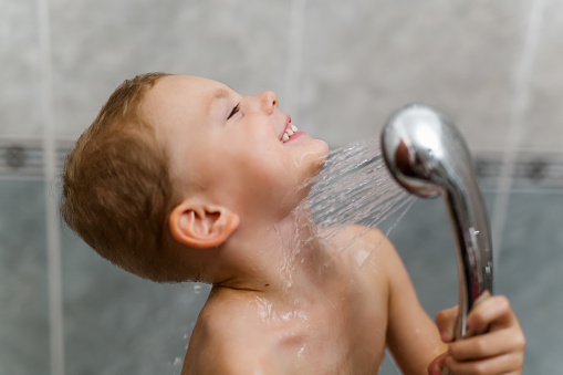 Cute happy smiling funny undressed boy child with blonde hair taking shower in bath with water indoor.
