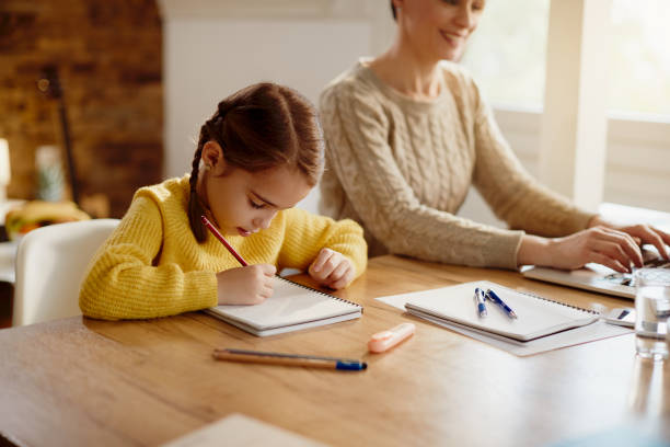 istock Little girl writing homework while he r mother is working on a computer at home. 1295838609