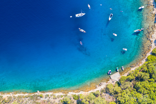Photo was captured from a drone and shows a view of the rocky beach and shallow waters of the Adriatic Sea (Jadransko More) on the Hvar Island, in Croatia. Several sailing boats, yachts and small vessels are moored or at anchor in this picturesque cove.