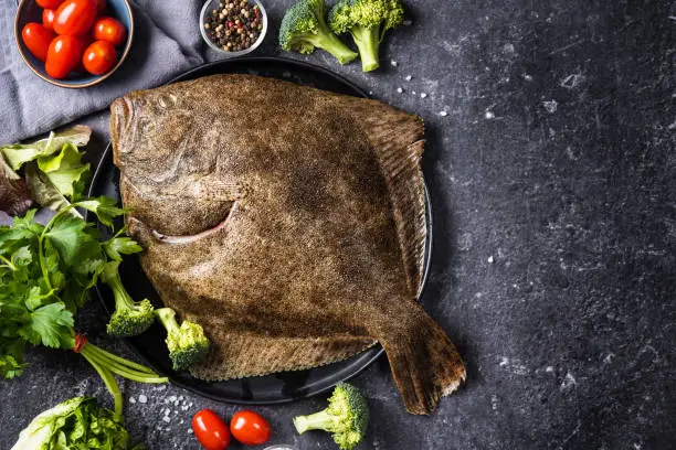 Top view of whole raw halibut fish rich of omega fats with vegetables on black background. Healthy for brain and mental clarity. Brain foods concept. Home cooking