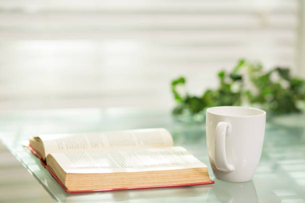 A  coffee mug sits on a glass top table near an open book.  Lush foliage off to one side.  In the background sunlight spills through the slats of the blinds covering the window.  Sunlight and reflections create a peaceful scene for an ideal morning break.