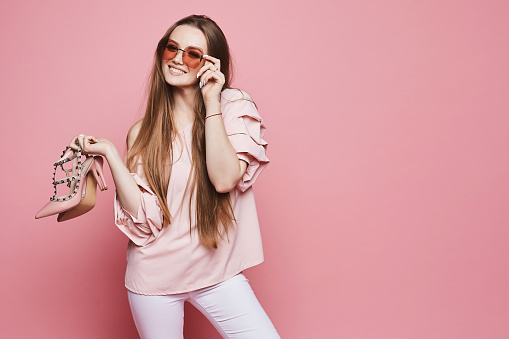 Chica modelo rubia feliz con una sonrisa brillante en blusa beige y gafas de sol rosa de moda sosteniendo zapatos elegantes y posando en el fondo rosa, aislado photo