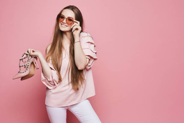 ragazza modello bionda felice con un sorriso lucido in camicetta beige e occhiali da sole rosa alla moda che tengono scarpe eleganti e posano sullo sfondo rosa, isolati - fashion foto e immagini stock