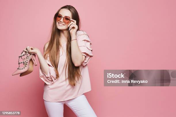 Ragazza Modello Bionda Felice Con Un Sorriso Lucido In Camicetta Beige E Occhiali Da Sole Rosa Alla Moda Che Tengono Scarpe Eleganti E Posano Sullo Sfondo Rosa Isolati - Fotografie stock e altre immagini di Moda