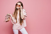 Happy blonde model girl with a shiny smile in beige blouse and fashionable pink sunglasses holding stylish shoes and posing at the pink background, isolated