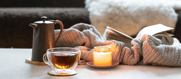 Glass cup of tea, teapot, candle and book with knitted element. The concept of home comfort and warmth.