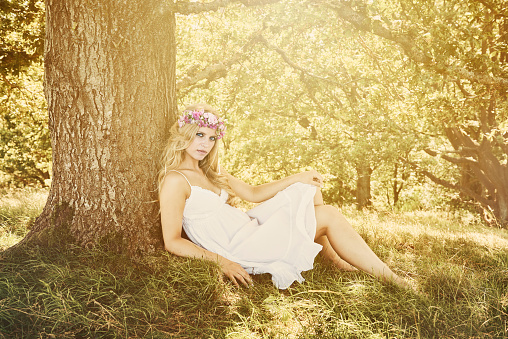 23 years old woman sitting in White summer dress under an oak tree. She is wearing some flowers in her hair. Blond Scandinavian girl looking at camera.