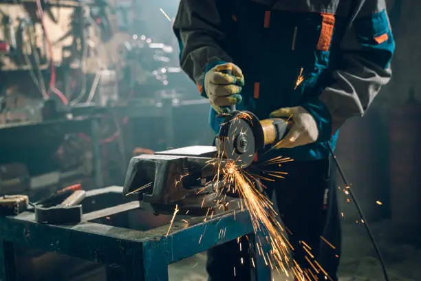 Photo of Locksmith in special clothes and goggles works in production. Metal processing with angle grinder. Sparks in metalworking