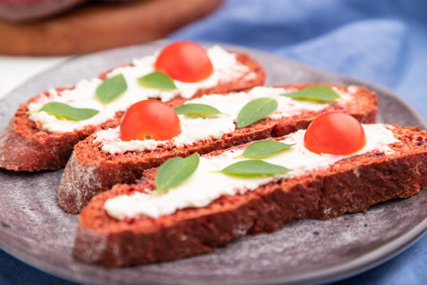 istock Red beet bread sandwiches with cream cheese and tomatoes on white concrete background and blue linen textile. Side view, selective focus, close up. 1295864747