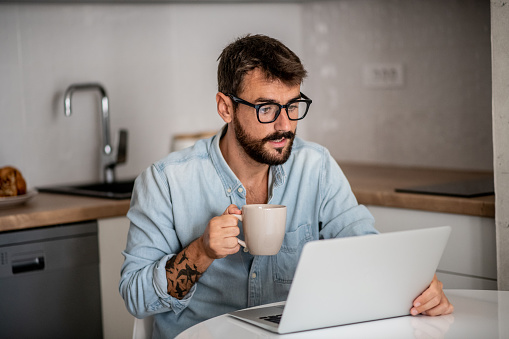 Young bearded hipster is sitting in coziness of his home and working online. He is sitting in kitchen at the table with laptop in front of him and drinking coffee.