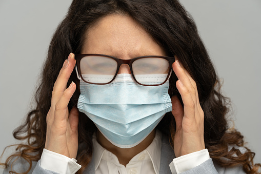 Business woman with foggy glasses from breath caused by wearing disposable mask on studio grey background. Protective measure during coronavirus pandemic.