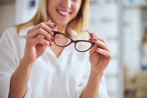 Cropped head portrait of adult beautiful lady in white shirt standing in room indoors while holding stylish set of eyeglasses in optometrist clinic
