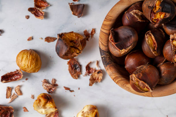 A wooden bowl of homemade oven roasted tsty chestnuts with shells and skins scattered on the marble table top. stock photo