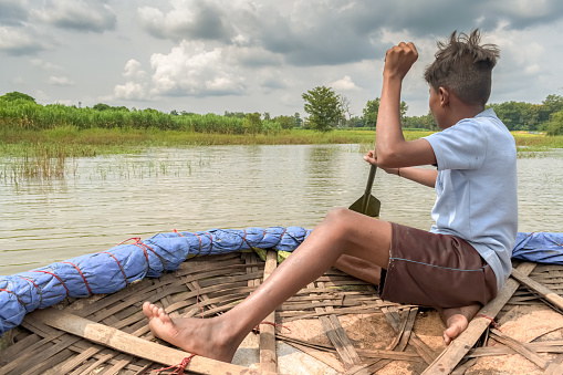 Amazing photo of a young boy rowing a tradition coracle made of bamboo woods, in river stream to cross the river in rural countryside background. He may be perceived as school going children who is struggling the basic necessities of proper transportation, or a young boy who is fishing for dinner, or as a hobby in leisure, or a tourist guide giving tourists a ride in traditional coracle. May be used for various other scenarios as well