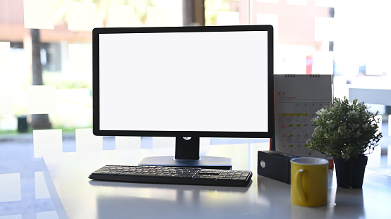 Large blank monitor with keyboard and office supplies on desk in eco-friendly office. Office cafeteria in the background. Render