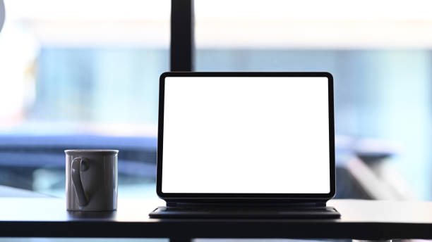 istock Front view of mock up tablet computer with blank screen and coffee cup on wooden table. 1295919101