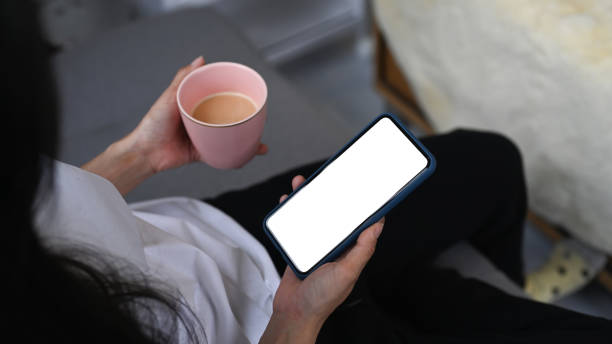 istock Close up view casual woman using smart phone and drinking coffee while sitting on couch at home. 1295919669