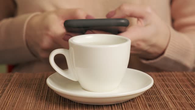 Female hands with mobile phone in hands and drinking coffee in white cup.