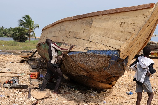 Aneho, Togo - November 20, 2019: Two men repair a traditional boat in Anheo, Togo, West Africa. All reusable parts are used again to avoid any waste. Each piece of recycled and re-useable wood is a precious raw material.