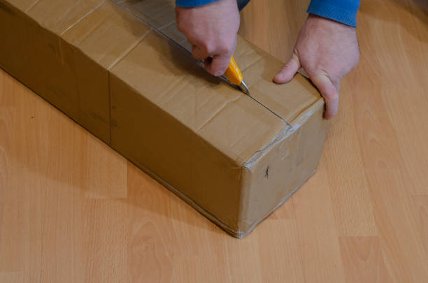 istock Man with yellow knife opens parcel in closed rectangular cardboard box on the floor. 1295936673