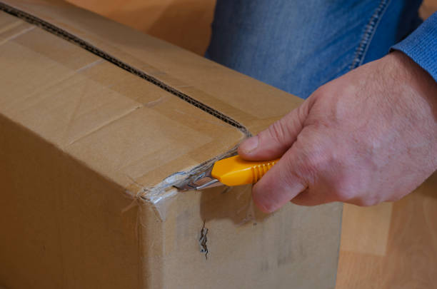 istock Man with yellow knife opens parcel in closed rectangular cardboard box on the floor. 1295936674