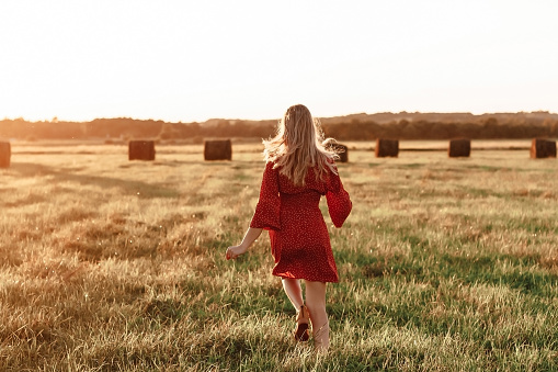 Young happy blonde in a red dress runs into a field at sunset