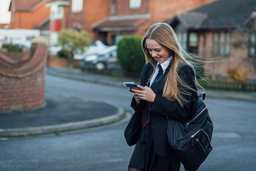 A three-quarter-length shot of a young caucasian girl wearing a school uniform is walking to school and using a smartphone.
