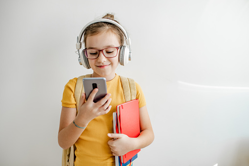 Portrait of a schoolgirl with backpack holding a notebook and mobile phone and standing in front of the white wall while having headphones on the head