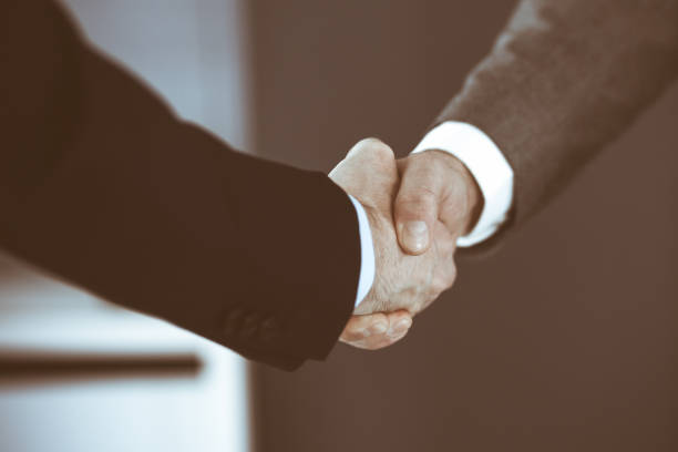 istock Business people shaking hands while standing with colleagues after meeting or negotiation, close-up. Group of unknown businessmen and women in modern office. Teamwork, partnership and handshake concept, toned picture 1295961956