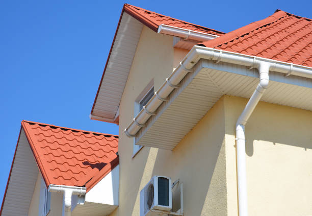 een stucwerk huis met een rood metalen dak, zolder en airconditioner outdoor unit met een close-up op een wit dak soffit boord en regen goot systeem met een downspout tegen blauwe hemel. - dakbalk stockfoto's en -beelden