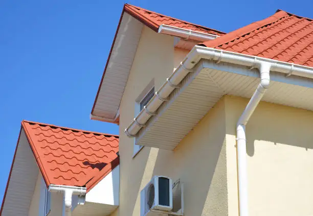 Photo of A stucco house with a red metal roof, attic and air conditioner outdoor unit with a close-up on a white roof soffit board and rain gutter system with a downspout against blue sky.