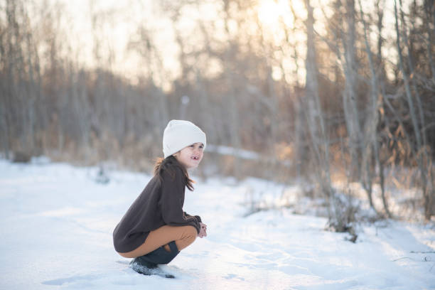 A Caucasian girl of elementary age is squatting on the snowy ground outdoors as she is patiently waiting to see if she'll see any birds in the woods.