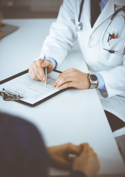 a doctor is talking to his patient, while sitting together at the desk in the cabinet in a hospital. physician using clipboard for filling up medication history records. perfect medical service in clinic. medicine and healthcare concepts - medicine cabinet medicine healthcare and medicine cabinet imagens e fotografias de stock