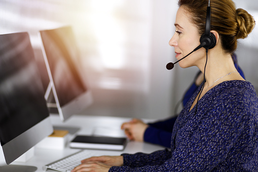 Two busineswomen have conversations with the clients by headsets, while sitting at the desk in a sunny modern office. Diverse people group in a call center. Telemarketing and customer service.