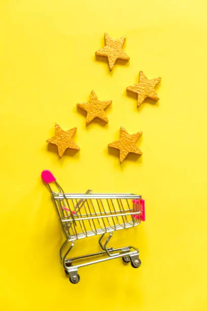 Photo of Simply flat lay design small supermarket grocery push cart for shopping with wheels and 5 gold stars rating isolated on yellow background. Retail consumer buying online assessment and review concept