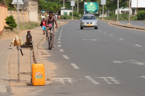 Aneho, Togo - November 20, 2019: Cyclist on the highway passing an illegal black market petrol station, which offers petrol in bottles and in a yellow container on the roadside. Location: N2 close to Church Paroisse Catholique Sacre-Coeur de Jesus Adjido in Aneho, Togo, West Africa.