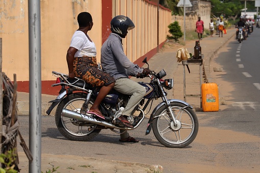 Aneho, Togo - November 20, 2019: Motorbike Taxi entering the highway. The male taxi driver uses a helmet. An illegal black market petrol station offers petrol in bottles and in a yellow container on the roadside. Location close to Church Paroisse Catholique Sacre-Coeur de Jesus Adjido in Aneho, Togo, West Africa.