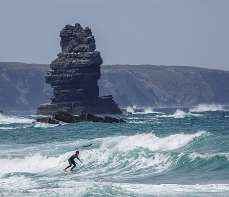 Arrifana, Algarve,Portugal- June 6, 2019: Lone surfer with towering rock in the background. Arrifana, Portugal.