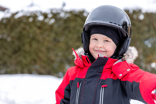 Close-up of a little redhead boy with green eyes, looking at camera. He is wearing a sports helmet while he is skiing outdoors in winter. He could also be ice-skating or snowboarding.