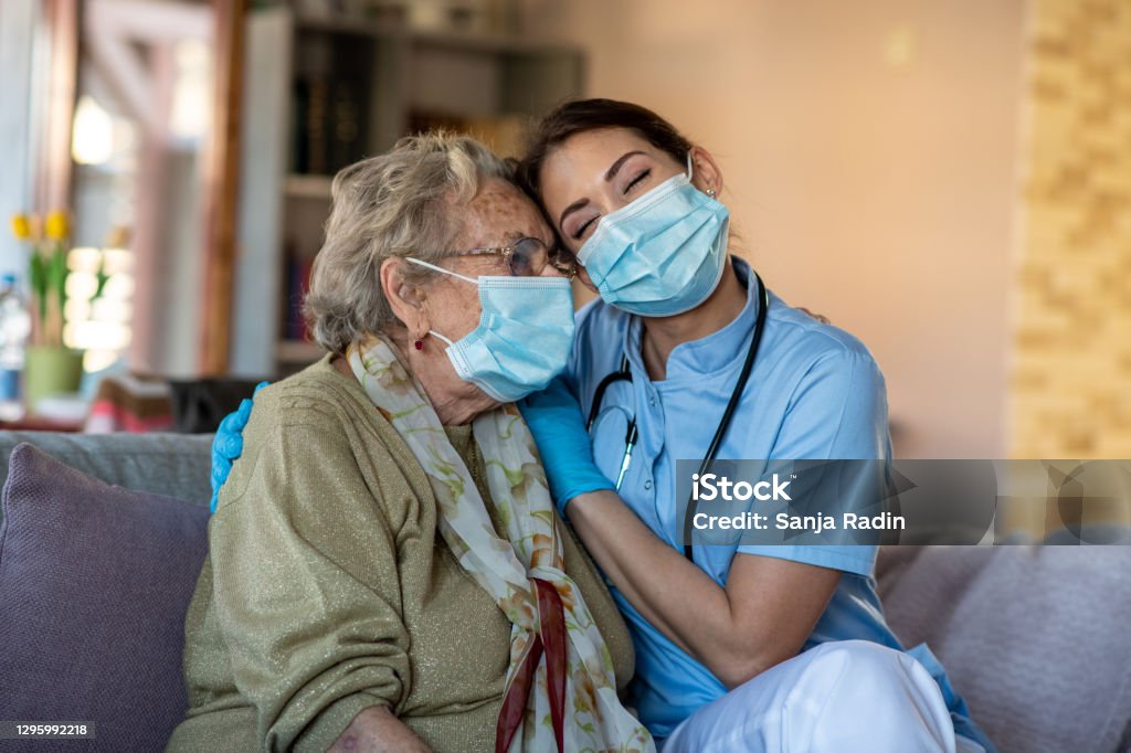 Nurse is hugging granny, helping her to get over COVID-19 crisis. Senior woman is sitting in her living room during home care visit. She is hugged by care specialist, consoled. Both of them have surgical masks on because of COVID-19. Home Caregiver Stock Photo