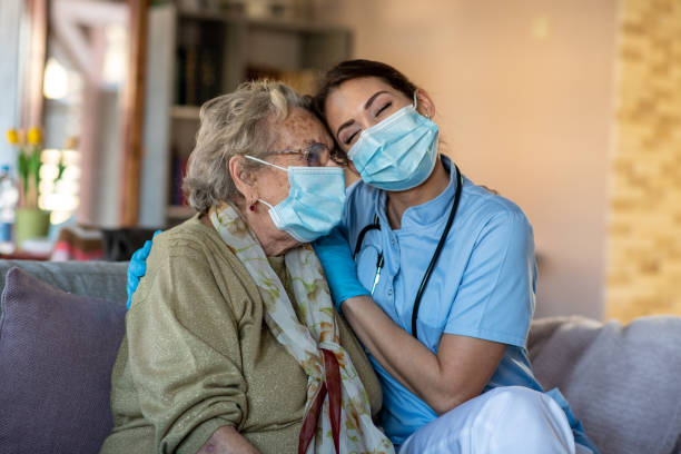 Nurse is hugging granny, helping her to get over COVID-19 crisis. Senior woman is sitting in her living room during home care visit. She is hugged by care specialist, consoled. Both of them have surgical masks on because of COVID-19. home caregiver photos stock pictures, royalty-free photos & images