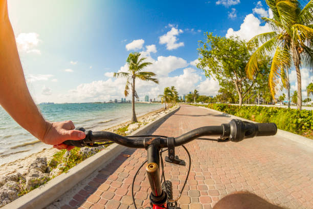 POV Point of view shot of a young latin sportsman barefoot wearing a blue swim shorts biking a bicycle in front of a paradise blue sea landscape over a brick pathway bicycle line at Hobie Island Beach Park in Key Biscayne, Miami Beach, Miami, South Florida, United States of America - USA. On background, a beautiful sunset view over a turquoise sea and white sand.

Shooting from a personal perspective in an exotic tropical beach travel holidays. Shotting location was next to Miami Seaquarium.