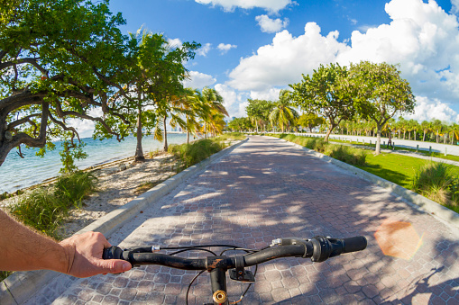 POV Point of view shot of a young latin sportsman barefoot wearing a blue swim shorts biking a bicycle in front of a paradise blue sea landscape over a brick pathway bicycle line at Hobie Island Beach Park in Key Biscayne, Miami Beach, Miami, South Florida, United States of America - USA. On background, a beautiful sunset view over a turquoise sea and white sand.\n\nShooting from a personal perspective in an exotic tropical beach travel holidays. Shotting location was next to Miami Seaquarium.