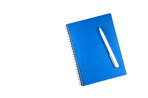 Closed blue notebook on a spiral for notes with a white ballpoint pen lying on the cover. Top view with copy space. Close-up on a white background.
