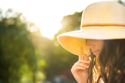 Close-up of a young woman holding the brim of her sun hat while standing alone outside in the summer