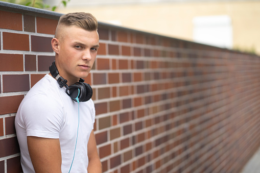 Teenage boy with headphones standing in front of a brick wall and looking in the camera